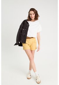Veste en denim noire, t-shirt blanc, shorts jaunes et baskets beiges avec des accents noirs. Le mannequin se tient avec une main sur la hanche, manches courtes.