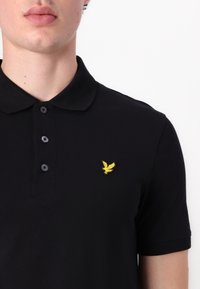 Homme portant un polo noir à manches courtes avec trois boutons et un petit logo d'oiseau jaune sur la poitrine gauche.