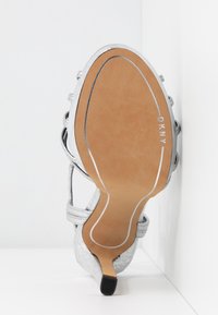 Sandale à talon haut argentée avec lanières, semelle marron clair, petite protection de talon, et marque "DKNY" sur la semelle, positionnée verticalement contre un coin blanc.