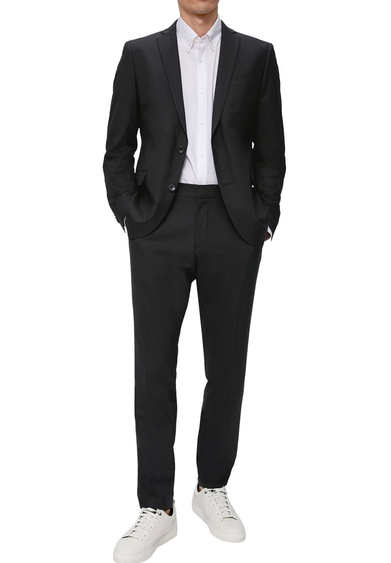 Schwarzer maßgeschneiderter Anzug mit einem taillierten Blazer und geraden Hosen, kombiniert mit einem weißen Hemd und weißen Sneakern.