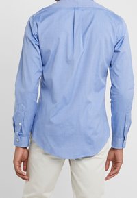 Muž v světle modré košili s dlouhým rukávem a světle béžových kalhotách, zobrazený zezadu na jednobarevném pozadí.