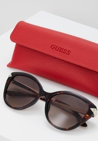 Ochelari de soare cu rame de tortoishell în formă de ochi de pisică, cu lentile întunecate și accente argintii. Însoțiți de un toc din piele roșie texturată, gravat cu "GUESS".