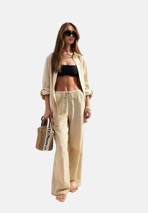 Crème-kleurige linnen outfit bestaande uit een losse, ongeknoopte shirt en broek met trekkoord. Zwart crop top en geweven tas met schouderband.
