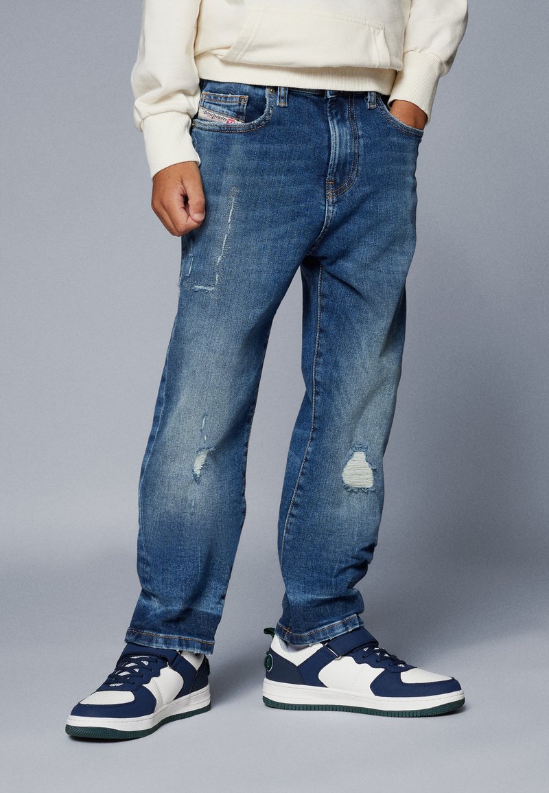 Jeans in denim di un blu medio, con dettagli abrasivi e una vestibilità comoda. Abbinati a sneakers bianche e blu navy, che evidenziano colori contrastanti.