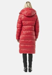 Roter Steppmantel mit Kapuze, mit horizontalem Steppmuster, glänzender Oberfläche und einem Logoaufnäher am linken Ärmel. Schwarze Hose und Stiefel.