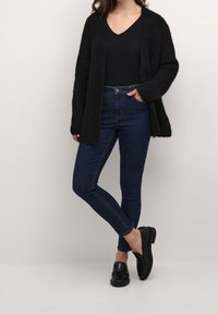 Musta pörröinen neuletakki, istuva musta paita, tummansiniset skinny-farkut ja mustat loaferit, jotka muodostavat kerroksellisen rentoon asuun erilaisia tekstuureja käyttäen.