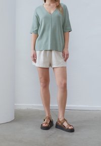 Maglione verde chiaro a V con maniche corte, abbinato a pantaloni corti crema con texture e sandali con piattaforma marroni dotati di fibbia.
