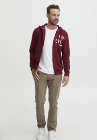 Burgunderfarget jakke med glidelås og hvit grafisk tekst, iført over en hvit t-skjorte, kombinert med khaki bukser og hvite sneakers.