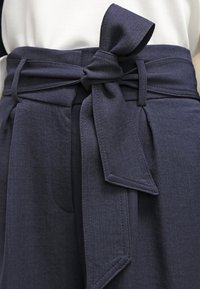 Námořnické vysoké kalhoty s širokým páskem zdobeným velkou mašlí. Tkanina má hladkou texturu a strukturovaný tvar s poutky na pásek.