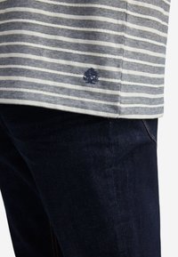 Pruhovaná šedá a bílá textilie s malým vyšitým logem stromu, spárovaná s tmavě modrými džíny s oranžovými švy.