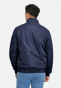 Chaqueta bomber azul marino con un exterior liso y brillante, puños y cinturilla elásticos. Presenta un cuello alto y sin herrajes visibles.