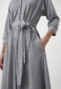 Robe grise à manches longues avec des boutons sur le devant, ceinture en tissu nouée à la taille, et main placée dans la poche droite.