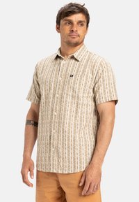Camisa de manga corta con botones en crema con rayas verticales marrones y patrones florales. Presenta un pequeño logo en el bolsillo del pecho.