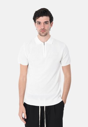 Giovane uomo con capelli ricci scuri che indossa una polo a maniche corte in maglia bianca con cerniera, abbinata a pantaloni neri con coulisse bianche.