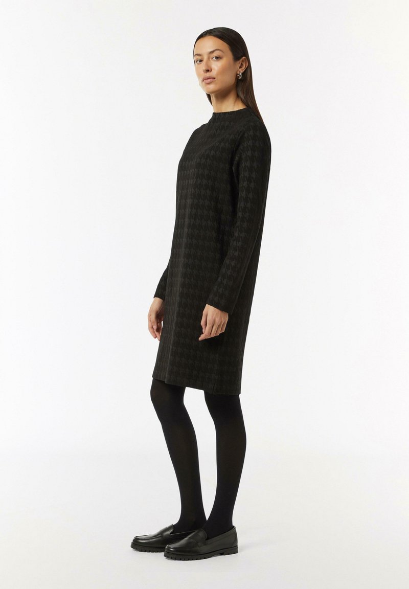 Schwarzes Kleid mit Hahnentrittmuster, runder Ausschnitt, lange Ärmel, knielang, kombiniert mit schwarzen Strumpfhosen und Slippern.