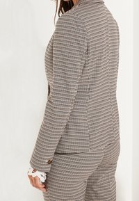 Blazer a scacchi in bianco e nero, vestibilità su misura, con colletto a punte, polsini con bottone e dettaglio di pizzo, e spalle strutturate.