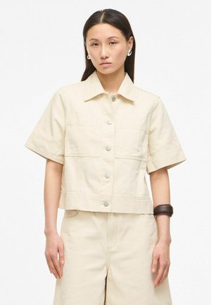 Femme aux cheveux raides et foncés porte une veste beige manches courtes boutonnée et un pantalon taille haute assorti, accessoirisés de boucles d'oreilles argentées et d'un bracelet foncé.