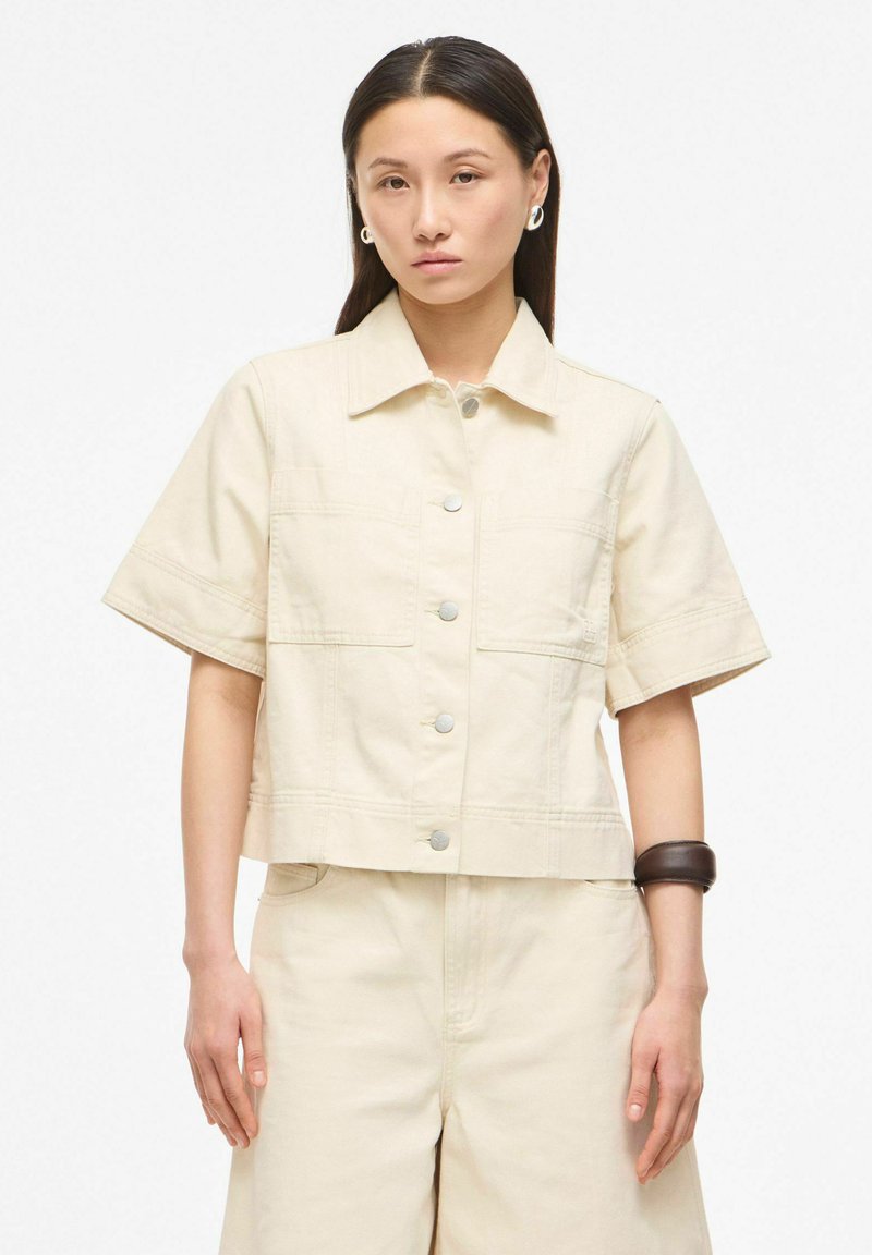 Femme aux cheveux raides et foncés porte une veste beige manches courtes boutonnée et un pantalon taille haute assorti, accessoirisés de boucles d'oreilles argentées et d'un bracelet foncé.