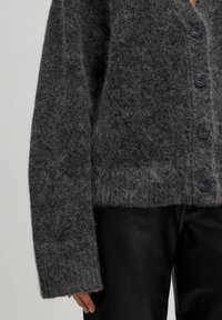 Grijze wollen cardigan met een zachte, fuzzy textuur, voorzien van een V-hals, verlaagde schouders en grote zwarte knopen. We gecombineerd met zwarte broeken.