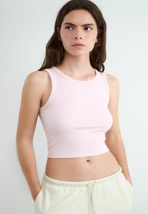 Ung kvinde med langt brunt hår iført en lys pink ærmeløs cropped top og cremefarvede bukser, stående med hænderne i lommerne.