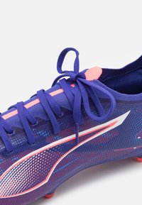 Botín de fútbol púrpura con acentos en rosa y blanco. Presenta una parte superior de malla transpirable, superficie texturizada y un sistema de cordones tradicional.