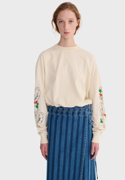 Žena u kremasti majici dugih rukava s uzorkom cvijeća na rukavima i plavoj prugastoj suknji na preklop, stoji ispred jednobojne pozadine.