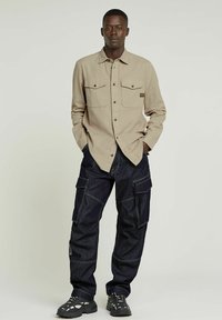 Beige långärmad skjorta med två bröstfickor, kombinerad med mörka denim cargo-byxor med flera fickor och kontrasterande sömmar. Svarta skor med utskärningar.