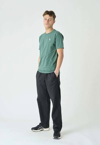 Zöld, pamut anyagú póló kerek nyakkivágással és kis hímzett logóval, fekete, bő fazonú nadrággal és fekete tornacipővel párosítva.