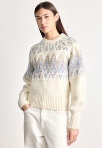 Jovem com cabelo escuro e comprido, vestindo um suéter creme com padrões geométricos azuis e bege e calças brancas, de pé com uma mão no bolso.