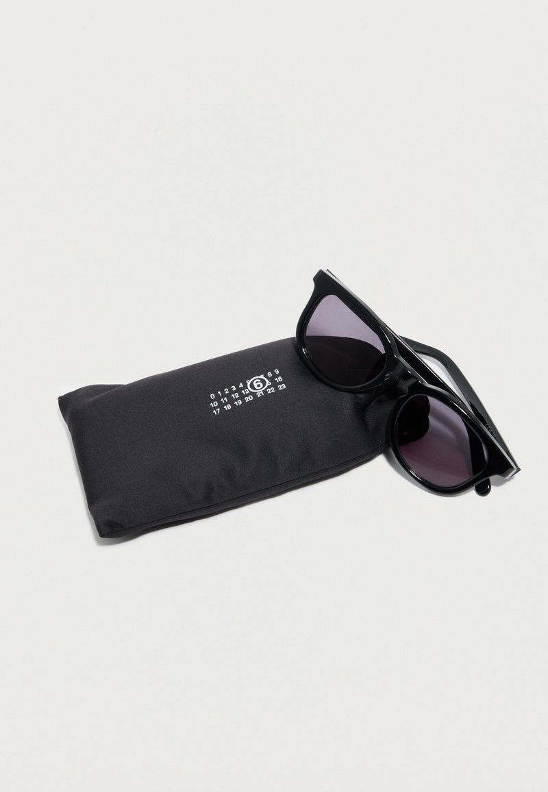 Gafas de sol negras con lentes oscuras descansando sobre una funda suave negra que presenta una cuadrícula de números impresa en blanco y un "6" rodeado en blanco sobre un fondo blanco.