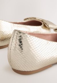 Aukso spalvos metalizuotos baleto bateliai su tekstūruotu, gyvatės odos pavidalo paviršiumi. Apval ūs pirštai ir mažytė kaspino detalė viršutinėje dalyje. Minkšta vidinė danga.