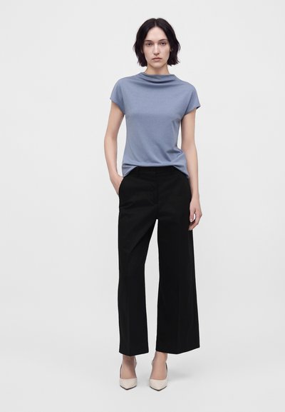 Femme aux cheveux courts et foncés, portant un haut bleu clair à manches courtes, un pantalon noir à jambes larges, et des talons pointus beiges, debout devant un fond uni.