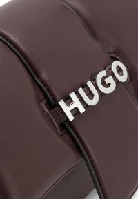 Burgundin värinen nahkalaukku, jossa on tyylikäs muotoilu. Laukussa on hopeinen "HUGO"-logo, musta ommel ja sileä pinta.