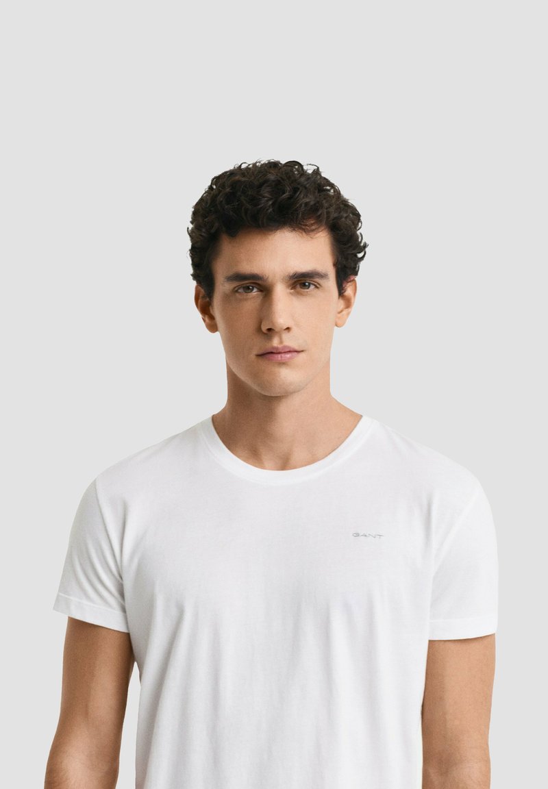 Giovane uomo con capelli ricci scuri che indossa una maglietta bianca semplice, di fronte a uno sfondo grigio chiaro.