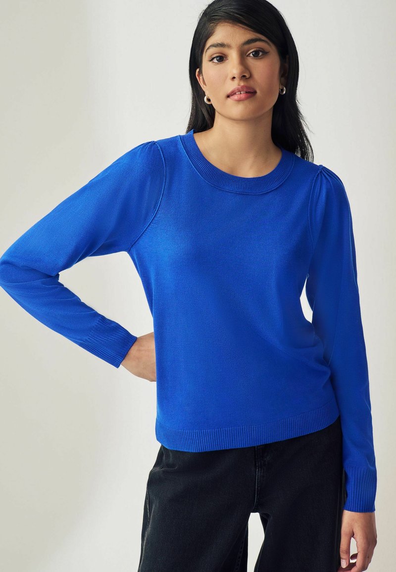 Pull en tricot bleu à manches longues, avec un col et un ourlet côtelés, présentant une légère épaule bouffante. Porté avec un pantalon noir.