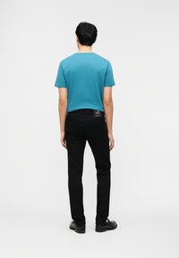 Άνδρας που στέκεται με την πλάτη προς τα εμπρός, φοράει γαλάζιο κοντομάνικο μπλουζάκι, μαύρα τζιν με εμφανή ετικέτα Replay, και μαύρα παπούτσια.