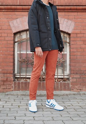 Persoon die een marineblauwe jas, een blauwgroene trui, roestkleurige broek en witte sneakers met blauwe strepen draagt, staand op het trottoir voor een bakstenen muur.
