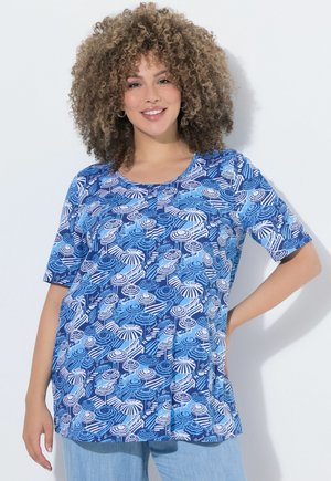 Kobieta o kręconych włosach, uśmiechająca się, ubrana w niebieski top z białym i niebieskim wzorem parasola, stojąca z jedną ręką na biodrze.