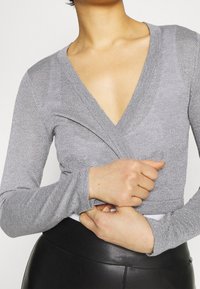 Maglietta grigia a maniche lunghe con scollatura a V profonda, tessuto testurizzato e design aderente. Sicura in vita, abbinata a un pantalone nero a vita alta.