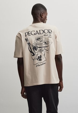 Mand iført beige t-shirt med sort illustration af mad og drikke samt teksten ""PEGADOR Under the Sun Vacation"" på ryggen.