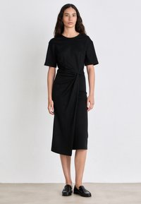 Vestito nero con un dettaglio annodato in vita, maniche corte e lunghezza midi. Indossato con scarpe slip-on nere. Tessuto dalla texture liscia.