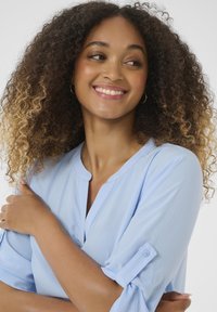 Blouse bleu clair avec un col rond et des manches boutonnées. Le tissu est lisse, avec un léger éclat et une coupe décontractée.