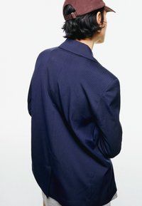 Blazer azul marino a rayas con una silueta estructurada, solapas con muesca y una abertura trasera, combinado con una gorra oscura y una camisa gris por debajo.