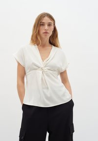Top bianco a maniche corte con dettaglio anteriore annodato, realizzato in tessuto morbido. Presenta un scollatura a V e un design drappeggiato, abbinato a pantaloni neri larghi.