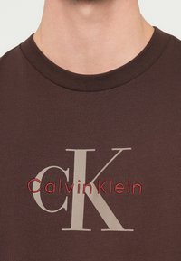 T-shirt castanha de algodão com gola redonda canelada, grande logótipo bege "CK" e texto vermelho "CalvinKlein" bordado na frente.