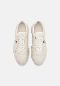 Pár krémových kožených tenisek s bílými tkaničkami, prošitým detailem a malým kovovým logem na boku na bílém pozadí.