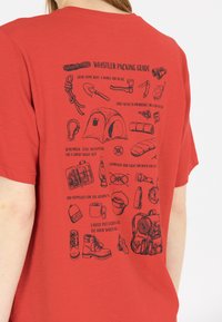 Punainen t-paita, jossa on musta kuvattu pakkausopas ulkoiluun, luettelee olennaiset varusteet ja tarvikkeet jäsennellyssä asettelussa.