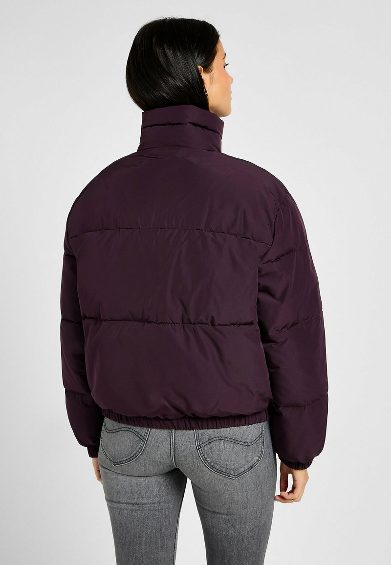 Paars pufferjack met een hoge kraag, voorzien van horizontale stiksels, elastische manchetten en een cropped ontwerp. Draagt over grijze jeans.