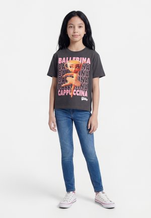 Fille aux longs cheveux noirs portant un t-shirt sombre avec un motif de ballerine cappuccino, un jean bleu et des baskets blanches sur fond blanc.