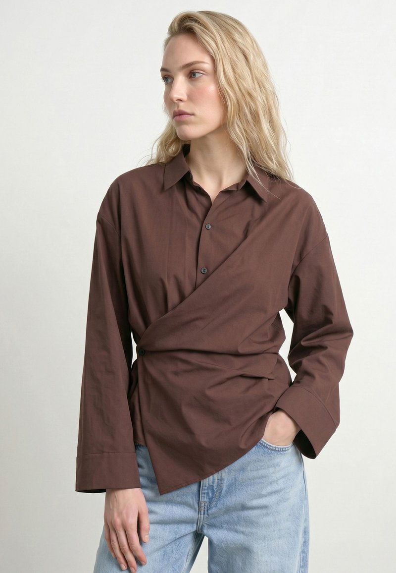 Femme blonde portant une chemise portefeuille marron à manches longues et un jean bleu clair, debout avec une main dans la poche, regardant vers la gauche.